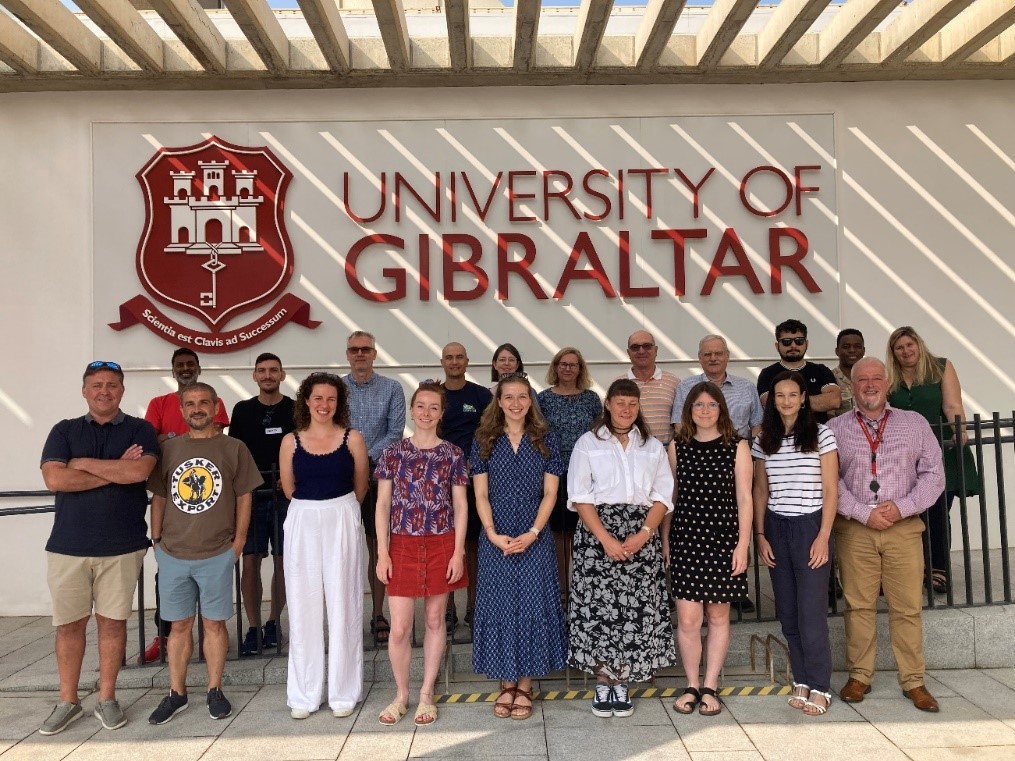 Group photo of 20 workshop participants in front of the University of Gibraltar sign