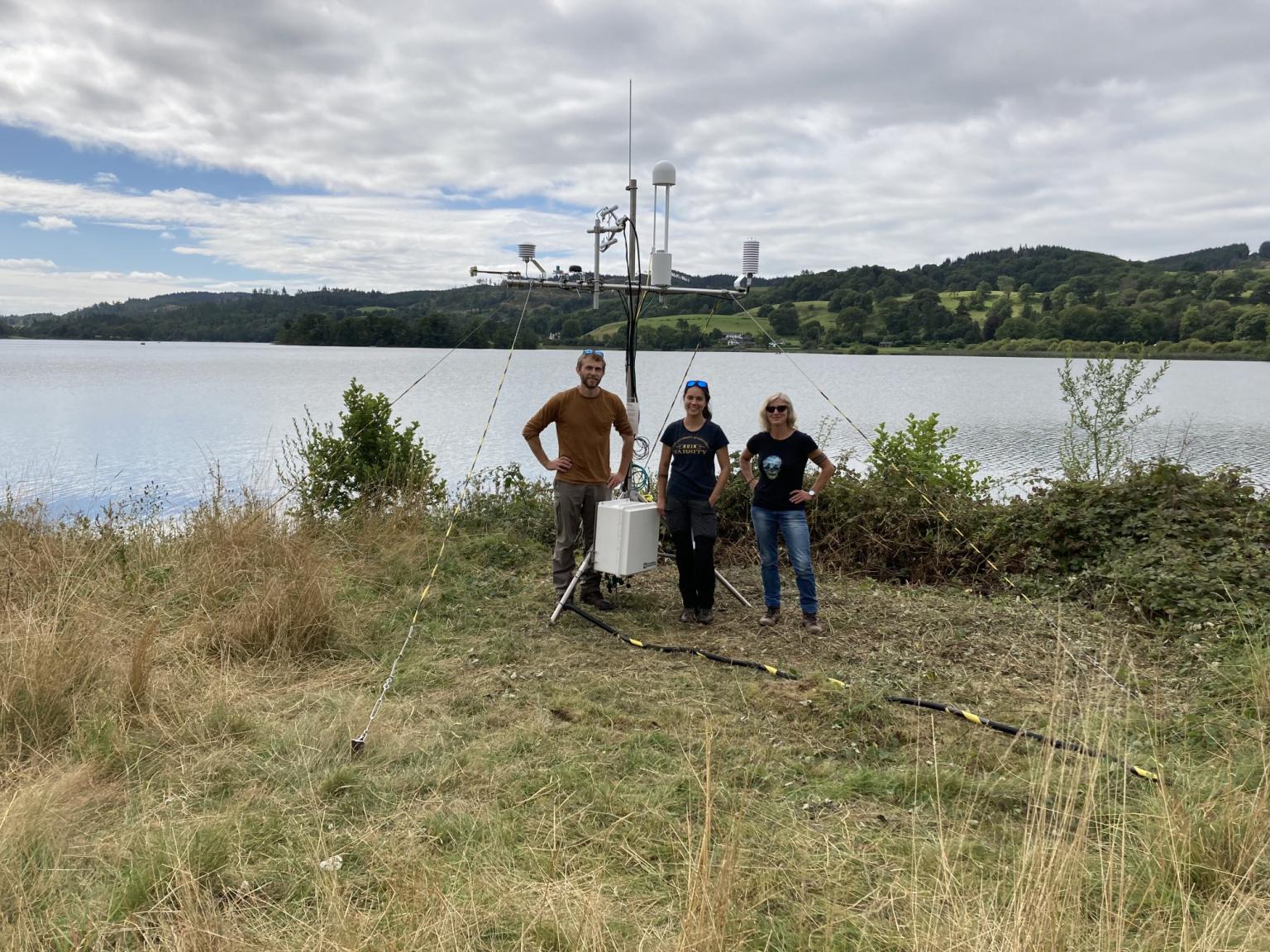 Three people standing beside flux measurement tower beside a lake