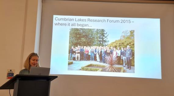 Ellie Mackay at the lectern with a slide showing a group photo and Cumbrian Lakes Research Forum 2015 - where it all began