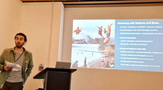 Presenter stands beside a lectern with a presentation slide in the background