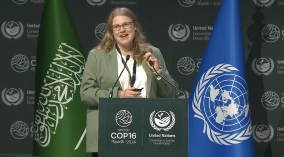 Lucy Barker behind lecturn with microphones presenting, UNCCD backdrop and green and blue flags behind