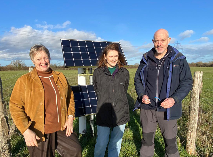 Three people stand in front of solar panels and scientific monitoring equipment in a field