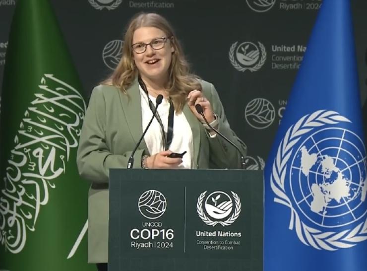 Lucy Barker behind lecturn with microphones presenting, UNCCD backdrop and green and blue flags behind