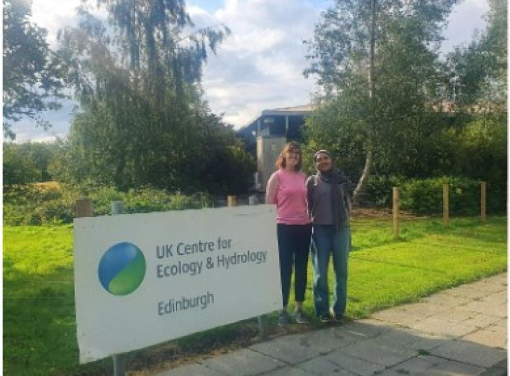 Two women standing alongside a UKCEH sign with grass and a building behind